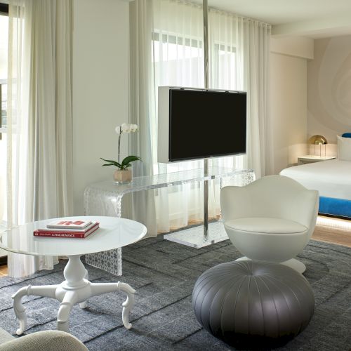 The image shows a modern hotel room with a bed, TV, chairs, round table, and large windows, featuring contemporary design elements.